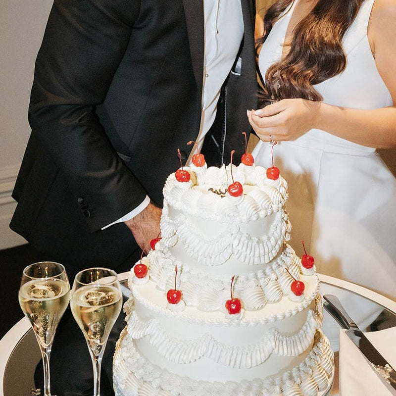 Bride and groom share a kiss beside a vintage-style wedding cake with piped details, cherries, and champagne glasses
